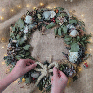 A close-up of hands adjusting a rope bow on a Coastal Christmas wreath of natural foliage.