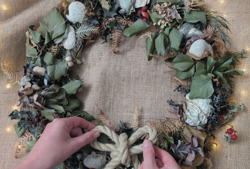 A close-up of hands adjusting a rope bow on a Coastal Christmas wreath of natural foliage.
