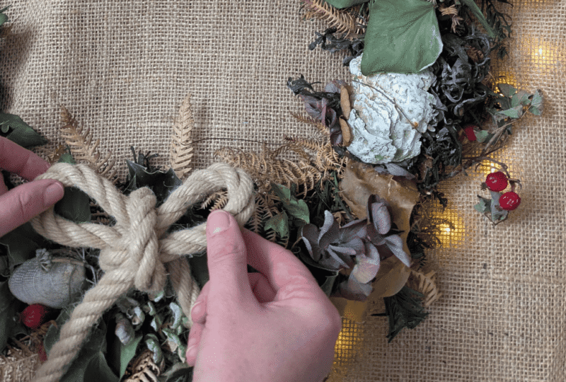 A close-up of hands adjusting a rope bow on a Coastal Christmas wreath of natural foliage.