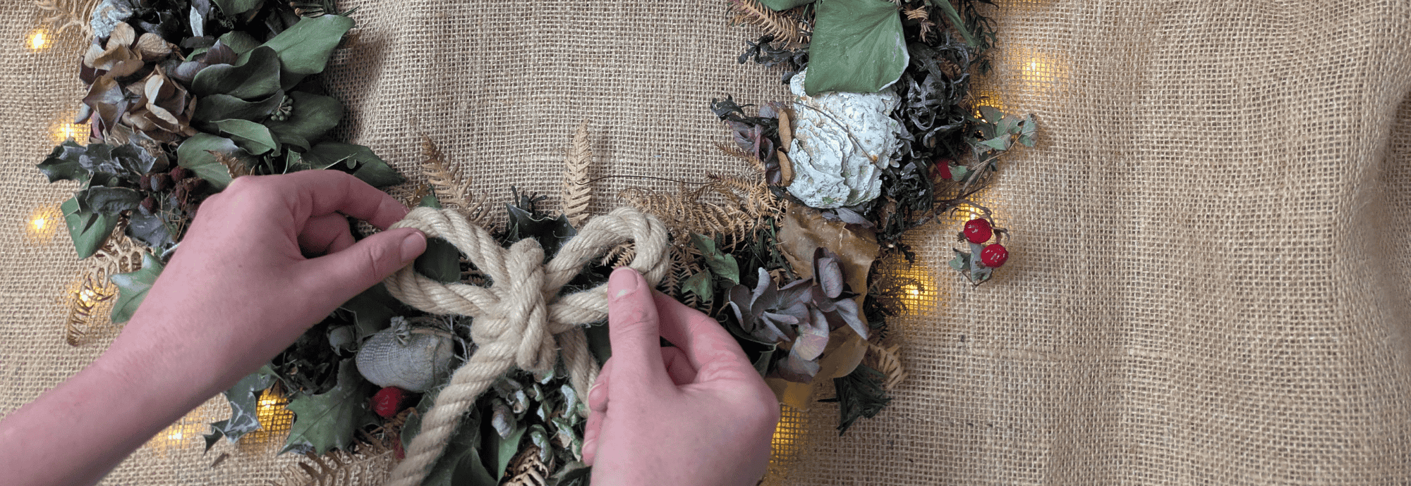 A close-up of hands adjusting a rope bow on a Coastal Christmas wreath of natural foliage.