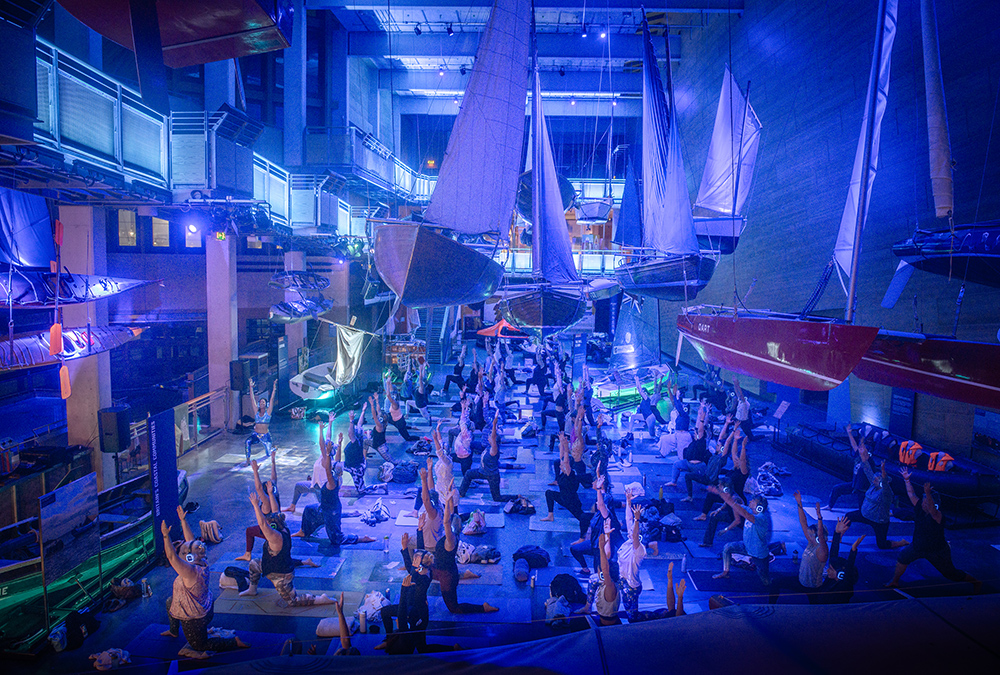 A group of people face a demonstrating yoga instructor, beside boat displays at NMMC at the Deep Blue Silent Disco Yoga event.