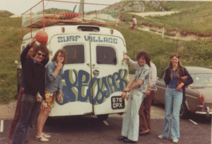 A group of people smiling at the camera stand around the back of a parked van, with surfboards on the roof-rack and the back doors painted with the words 'Surf Village Skewjack'.