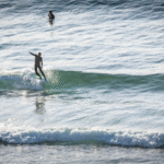 Surfer Sam Bleakley stands on a surfboard, riding a wave with one arm in the air, the other arm at his side.