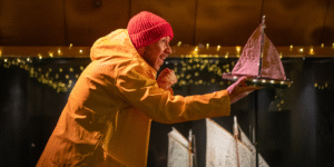 A performer in the Nadelik Christmas show is dressed in a red hat and yellow fisherman's anorak, holding a model boat with a backdrop of fairy lights.