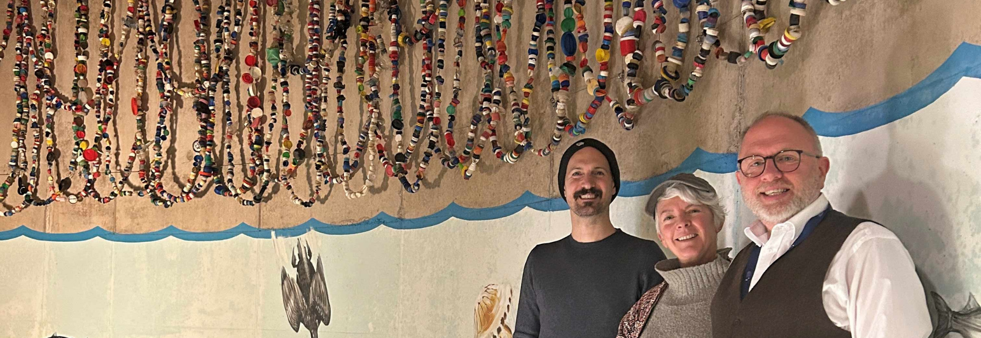 Three people stand in front of a wall painted with underwater scenes, above them is a hanging display of colourful bottle tops.