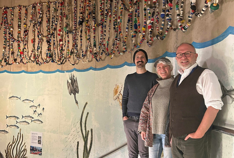 Three people stand in front of a wall painted with underwater scenes, above them is a hanging display of colourful bottle tops.
