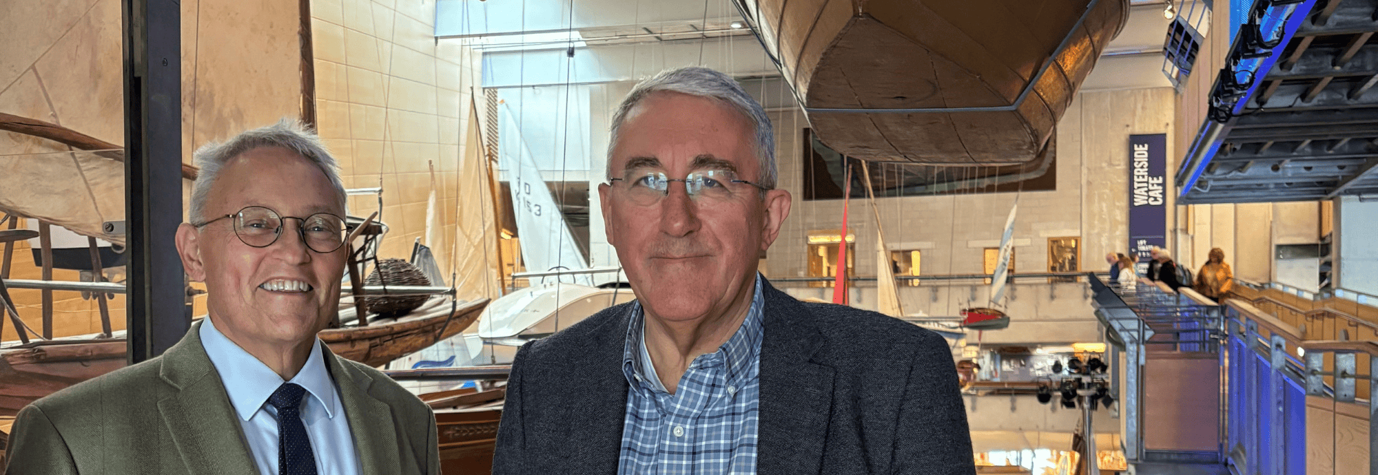 Two men, NMMC Director Richard Doughty and NMMC Board Chair Dr Patrick Newberry, stand side-by-side in the Boat Hall of National Maritime Museum Cornwall.
