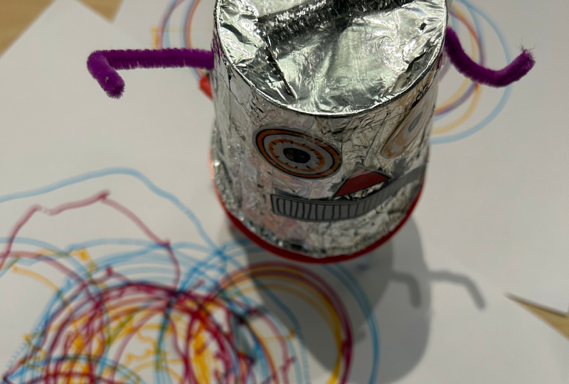 A tinfoil covered cup-shaped scribblebot, with paper eyes and mouth, stands on a colourful drawing it has just created.
