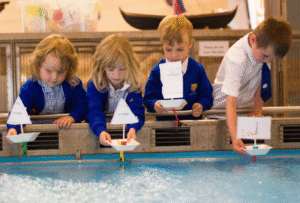 Four uniformed school children each hold a homemade paper boat over the edge of the boat pool at National Maritime Museum Cornwall, ready to let go and watch them float.