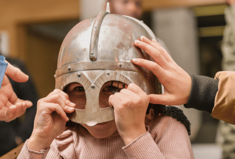A child holds a metal viking helment on their head, their eyes show through a frame.