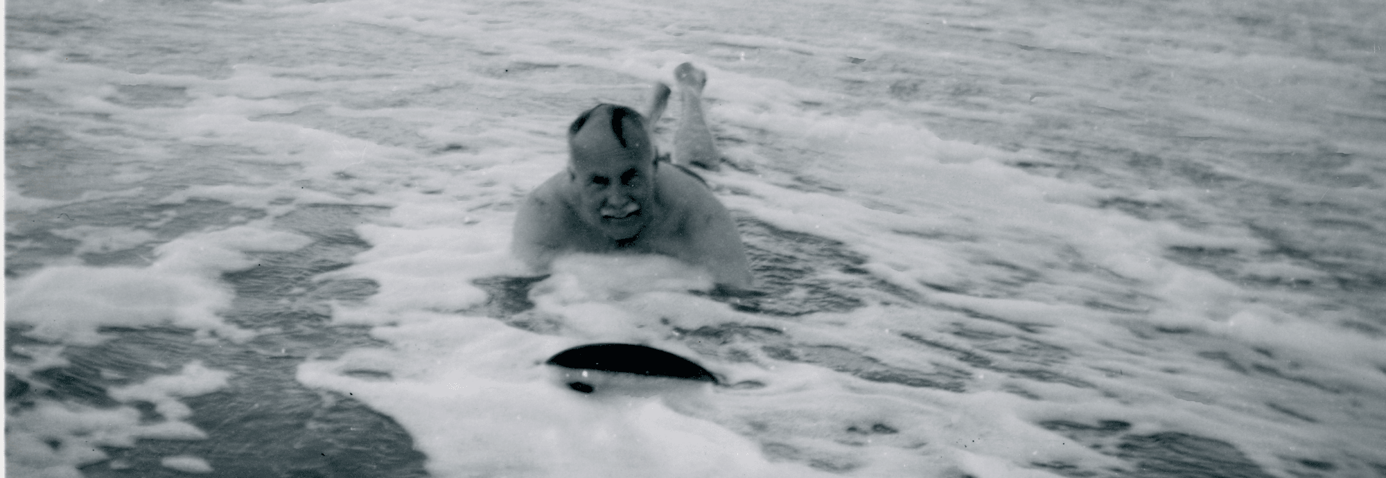 A black and white photograph of a man smiling as he is riding a wooden board into shallow water towards the camera, figures can be seen in the distance standing in shallow water.