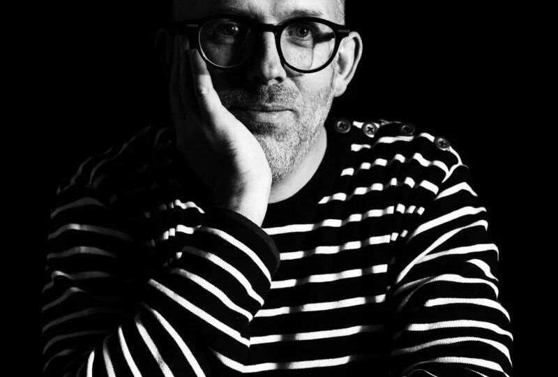 Black and white portrait photograph showing author Wyl Menmuir wearing a striped shirt.