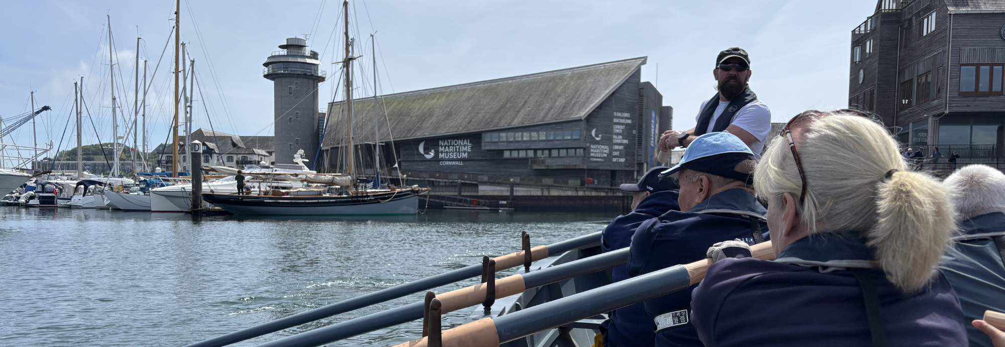 Devoran Gig Club Rowers Hold the Oars of the Museum's WW2 Surfboat replica as they row through the harbour, with National Maritime Museum Cornwall in the background.