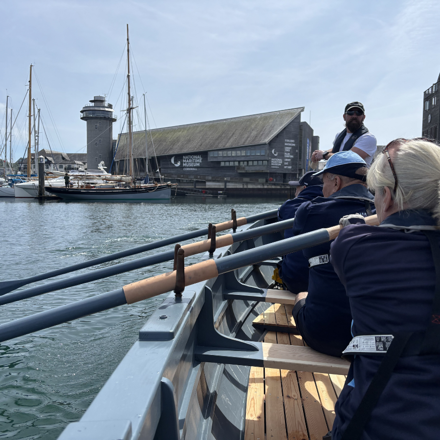 Devoran Gig Club Rowers Hold the Oars of the Museum's WW2 Surfboat replica as they row through the harbour, with National Maritime Museum Cornwall in the background.