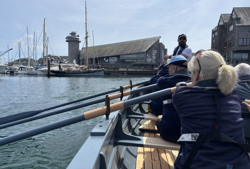 Devoran Gig Club Rowers Hold the Oars of the Museum's WW2 Surfboat replica as they row through the harbour, with National Maritime Museum Cornwall in the background.