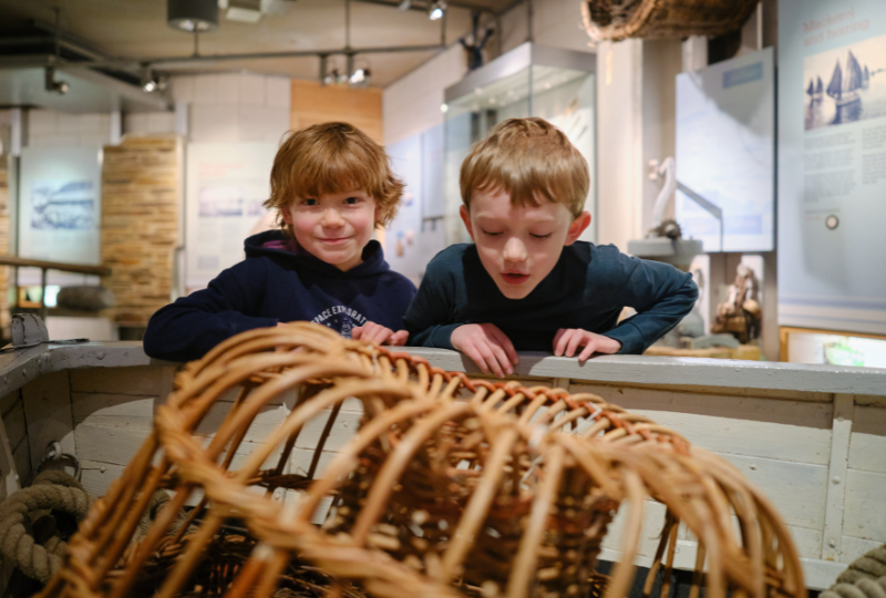 Two children peer over the side of a display boat to take a closer look at the willow lobster pots inside at National Maritime Museum Cornwall.