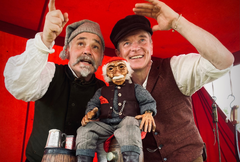 Two actors from Rattlebox Theatre wear waistcoats and hats while standing beside a puppet of an old man, also wearing a waistcoast and striped trousers, with a woolen hat and fluffy white moustache.