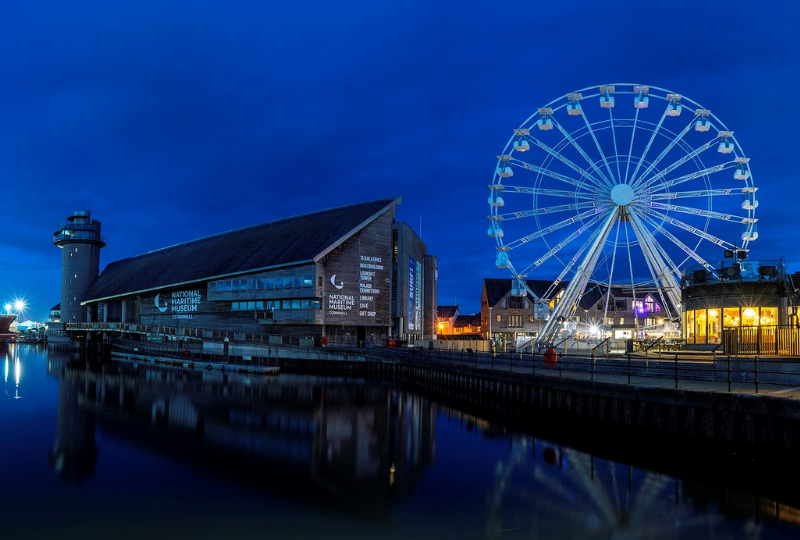 A large Observation Wheel stands beside the National Maritime Museum Cornwall and nearby buildings at night, the water of Falmouth harbour in the foreground.